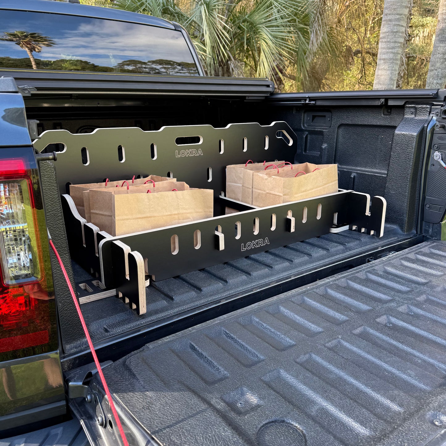 A black LOKRA truck bed organizer installed in the bed of a pickup truck, holding two brown paper bags. The truck is parked outdoors with palm trees visible in the background.