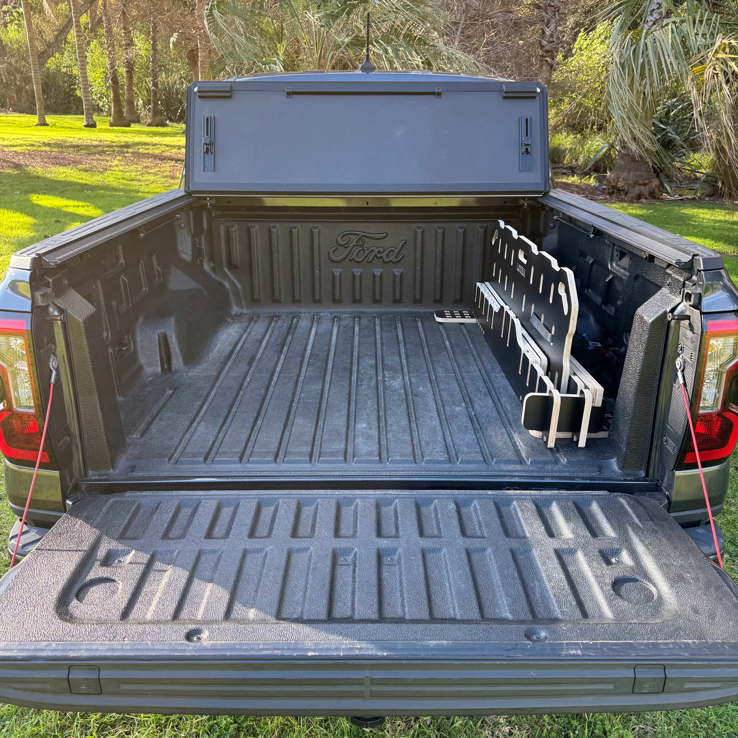 A black Ford truck bed liner installed in the bed of a pickup truck, shown outdoors with grass and trees in the background. The liner features a textured floor, a raised tailgate lip, and a side wall with a Ford logo embossed on the back panel.