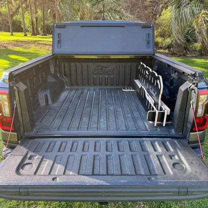 A black Ford truck bed liner installed in the bed of a pickup truck, shown outdoors with grass and trees in the background. The liner features a textured floor, a raised tailgate lip, and a side wall with a Ford logo embossed on the back panel.
