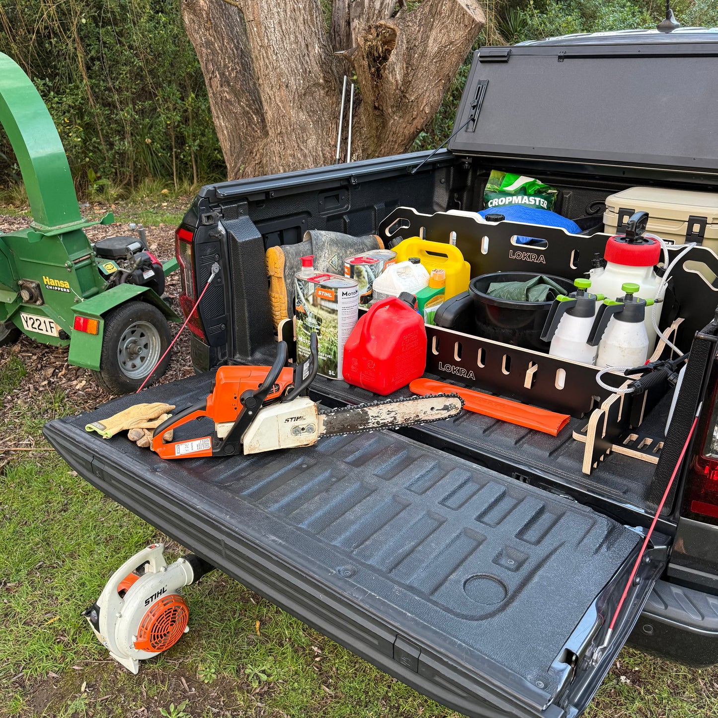 A black LOKRA UTE TRAY DIVIDER STORAGE filled with outdoor tools and equipment, including a chainsaw, orange gas canisters, spray bottles, and a leaf blower, set in a backyard with a green wood chipper and tree trunk in the background.